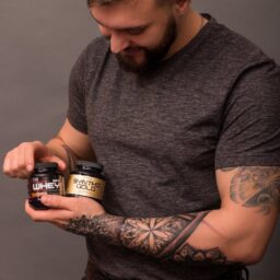Bearded man with arm tattoos holding protein supplement bottles in a studio setting.