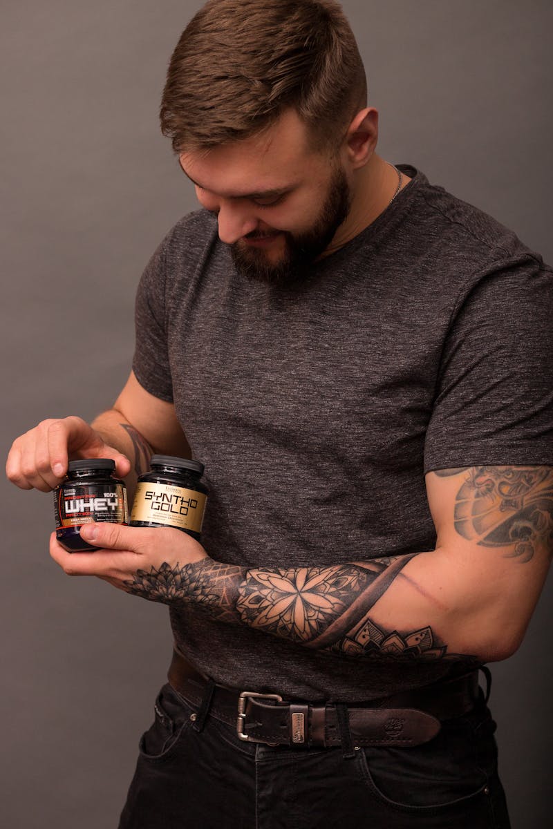 Bearded man with arm tattoos holding protein supplement bottles in a studio setting.